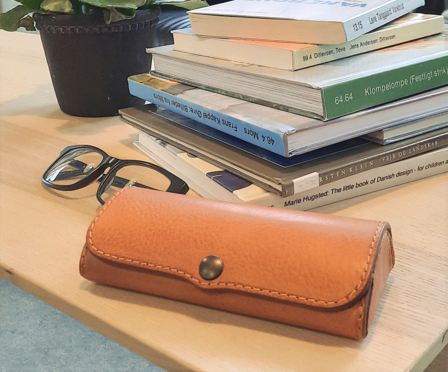 Light brown leather eyeglass case, seen from the side, resting on a table with books and glasses.